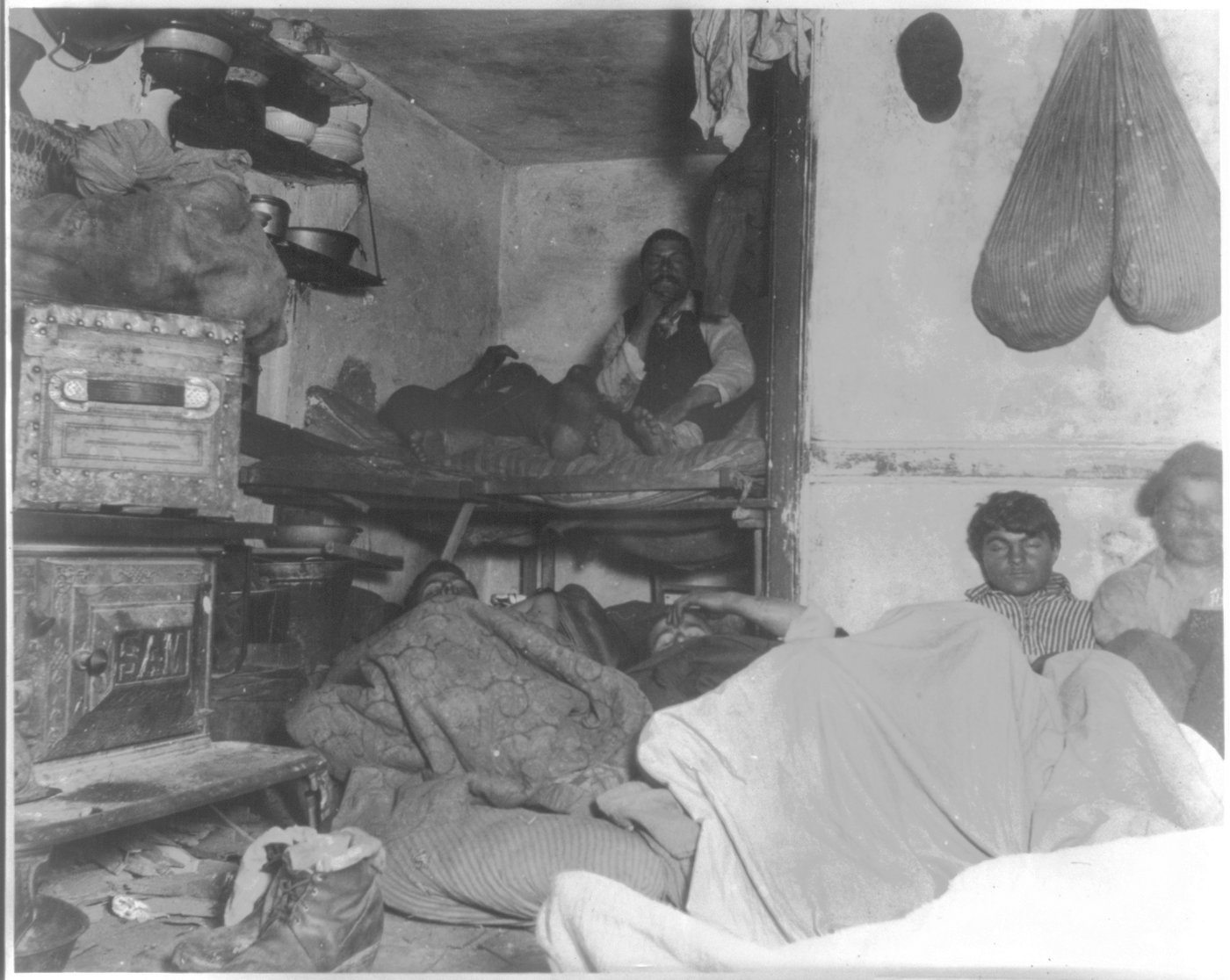 A black-and-white flash photograph of men crowded onto a bunk and the floor of a small, dim tenement lodging room, with cookware, bundled bedding and a stove crammed into the same space.