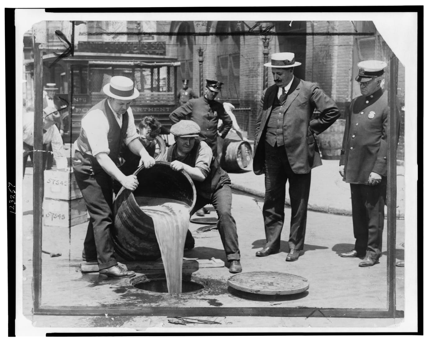 Men in suits and hats pour liquor from confiscated barrels into a New York street sewer during Prohibition.