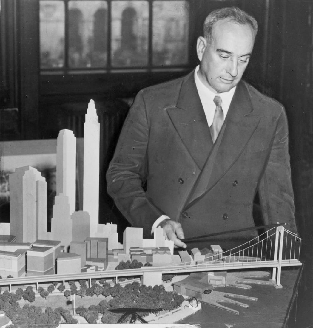 Robert Moses in a suit standing beside a large tabletop model of a proposed New York bridge in 1939.