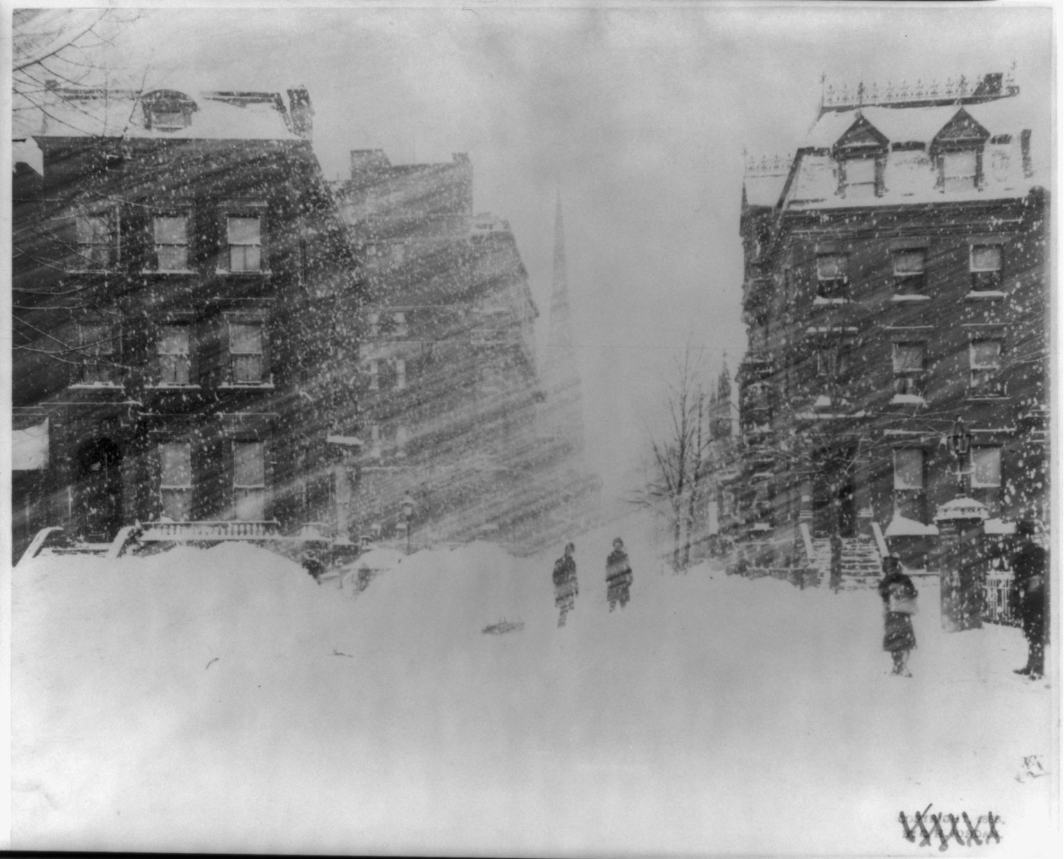 A photograph of a New York street in driving snow during the 1888 blizzard, with a few pedestrians between snow-covered rowhouses and drifts shoulder high.