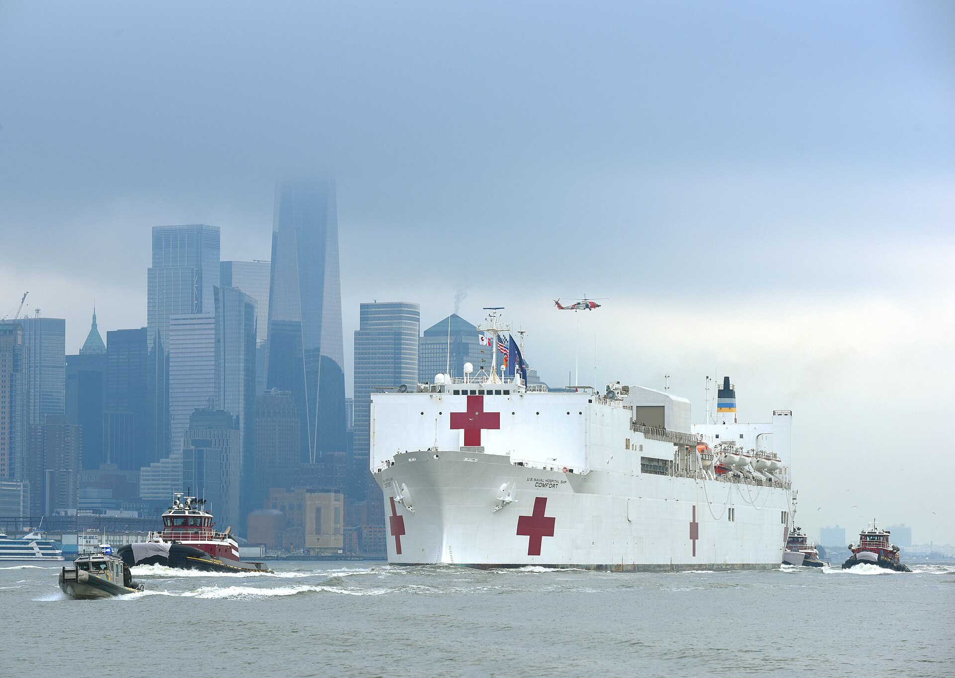 The white Navy hospital ship USNS Comfort, marked with red crosses, entering New York Harbor with tugboats and a police boat, the Lower Manhattan skyline shrouded in fog behind it.