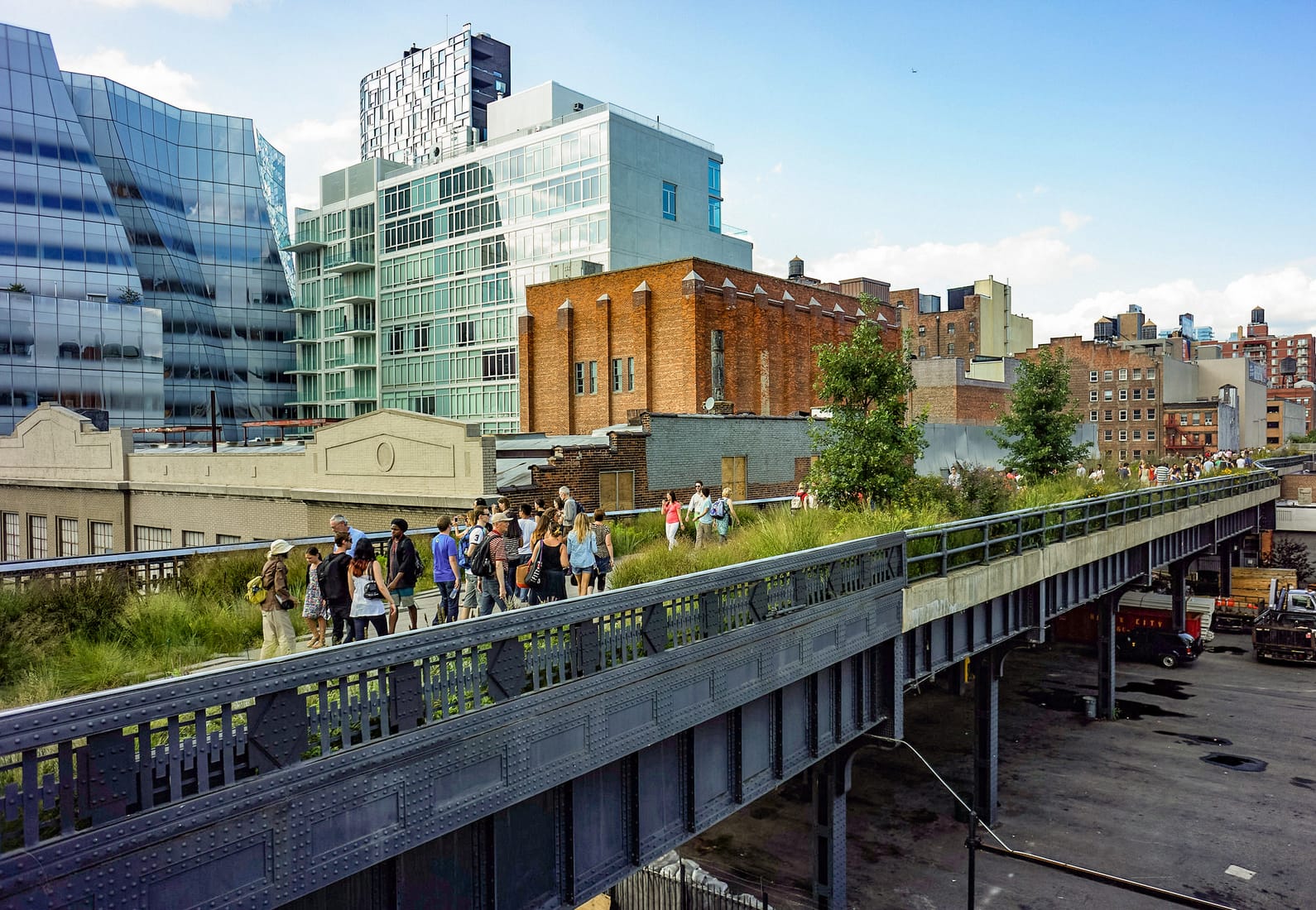 People walking along a planted path on the elevated High Line park in Manhattan, with modern glass buildings rising around it.